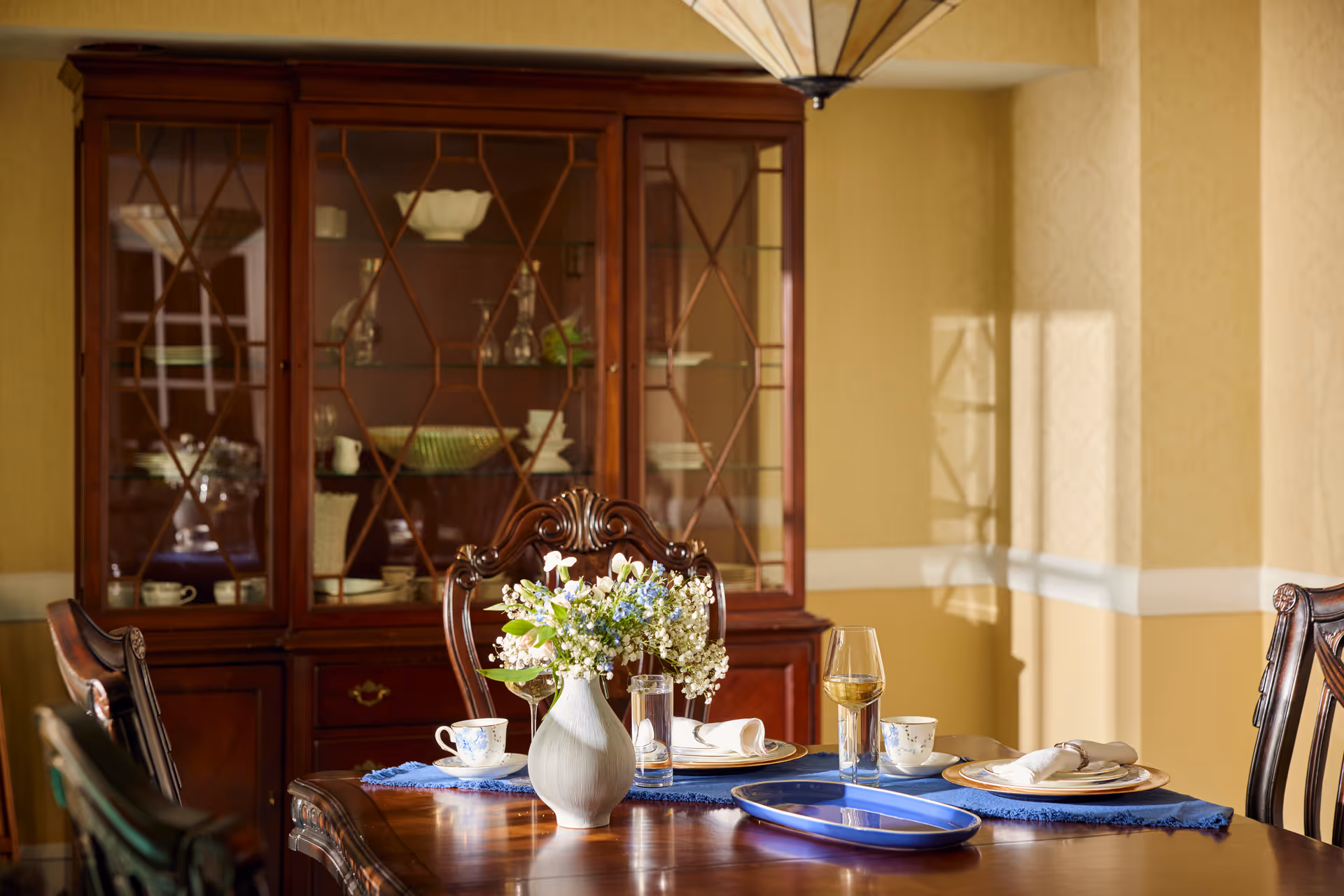A dining room with a wooden table set for a meal, featuring a white vase with flowers, teacups, plates, napkins, and a glass of white wine. Behind the table is a wooden china cabinet with glass doors displaying dishes and glassware. The room has warm yellow walls and soft natural light coming through a window.