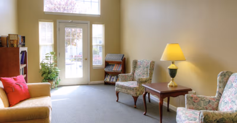 Sunlit common sitting area with upholstered armchairs and a sofa arranged around a side table with a lamp, a small bookcase, and a glass door leading outside.