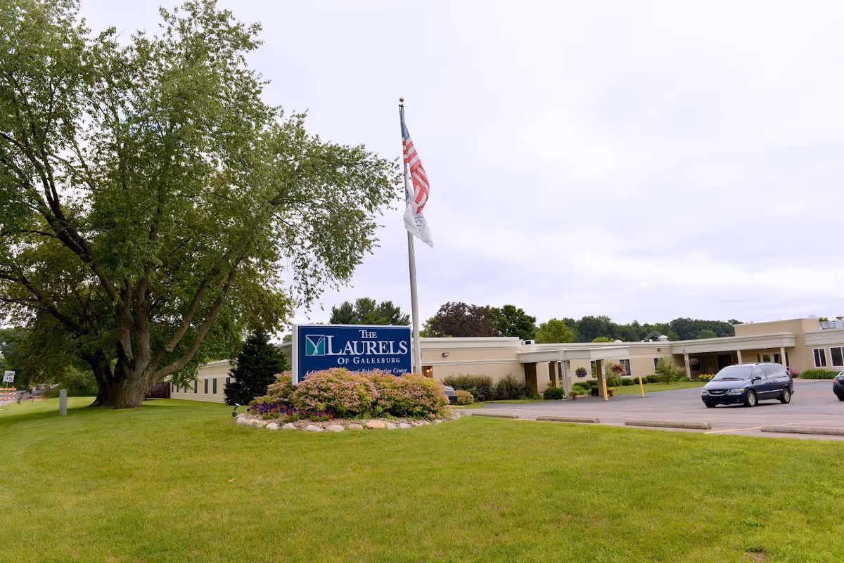 Front exterior of The Laurels of Galesburg senior living facility with a sign, flagpole, lawn, and parked cars.