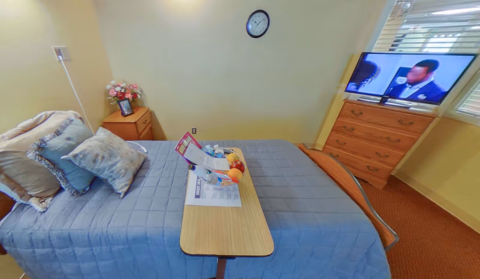 A single bed in a private room with pillows and an overbed table holding snacks and papers, a bedside table with flowers, a dresser with a TV, and a wall clock.