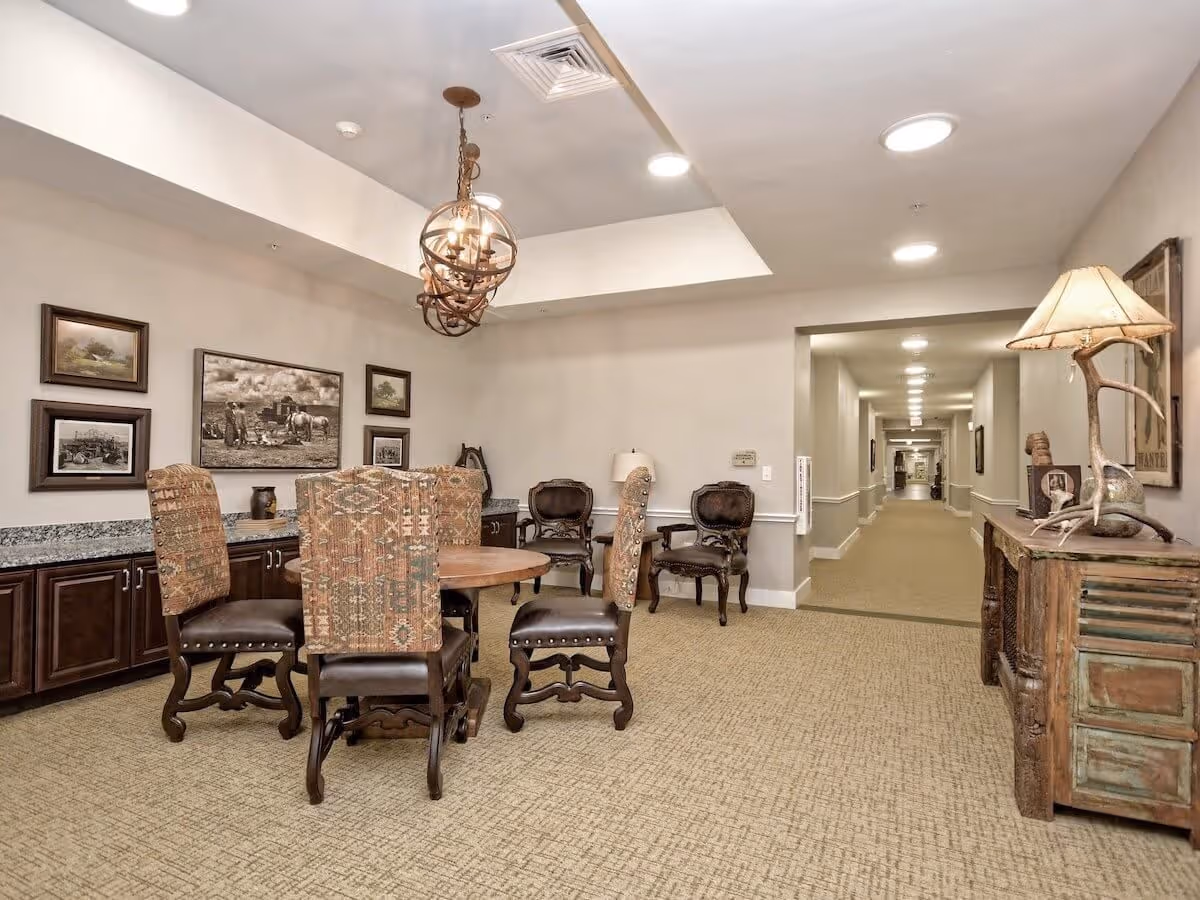 A furnished common dining area with a round wooden table, upholstered chairs, framed artwork on the wall, and a long hallway leading into the facility.