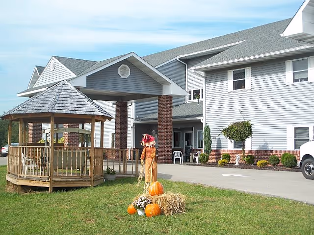 Exterior view of a senior living facility named Assisted Living At Evergreen featuring a two-story building with gray siding and brick accents. In front of the building is a wooden gazebo with chairs inside, a small scarecrow decoration, pumpkins, and hay bales on a grassy lawn under a blue sky.