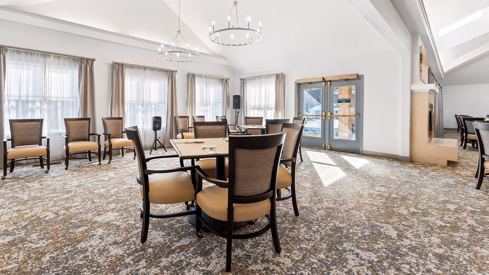 Bright dining room with multiple tables and chairs arranged on a patterned carpet. Large windows with sheer curtains allow natural light to fill the space. Two modern chandeliers hang from the ceiling, and double glass doors lead outside. Additional seating is visible along the walls and in an adjacent room.