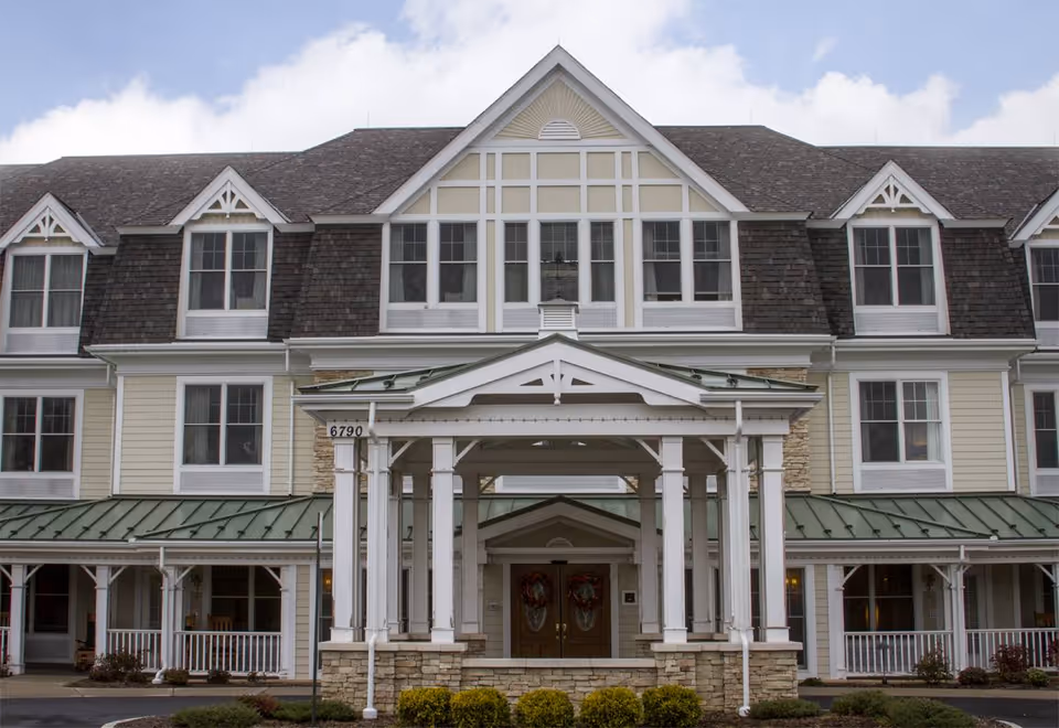 Front exterior view of a large, multi-story senior living facility building with beige siding, white trim, and a green metal roof over the entrance. The entrance features a covered porch supported by white columns and stone accents. The building has multiple windows and dormers on the roof, with a partly cloudy sky in the background.