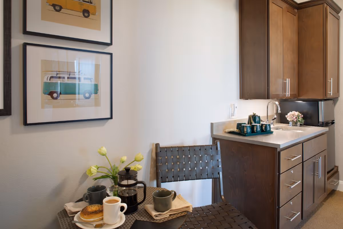A small kitchenette area with wooden cabinets, a countertop with a sink, a small refrigerator, and a tray with a teapot and cups. In front of the kitchenette is a small round table with a coffee pot, two mugs, a plate with a bagel, and a vase with yellow tulips. Two framed pictures of vintage vans hang on the wall above the table and chair.