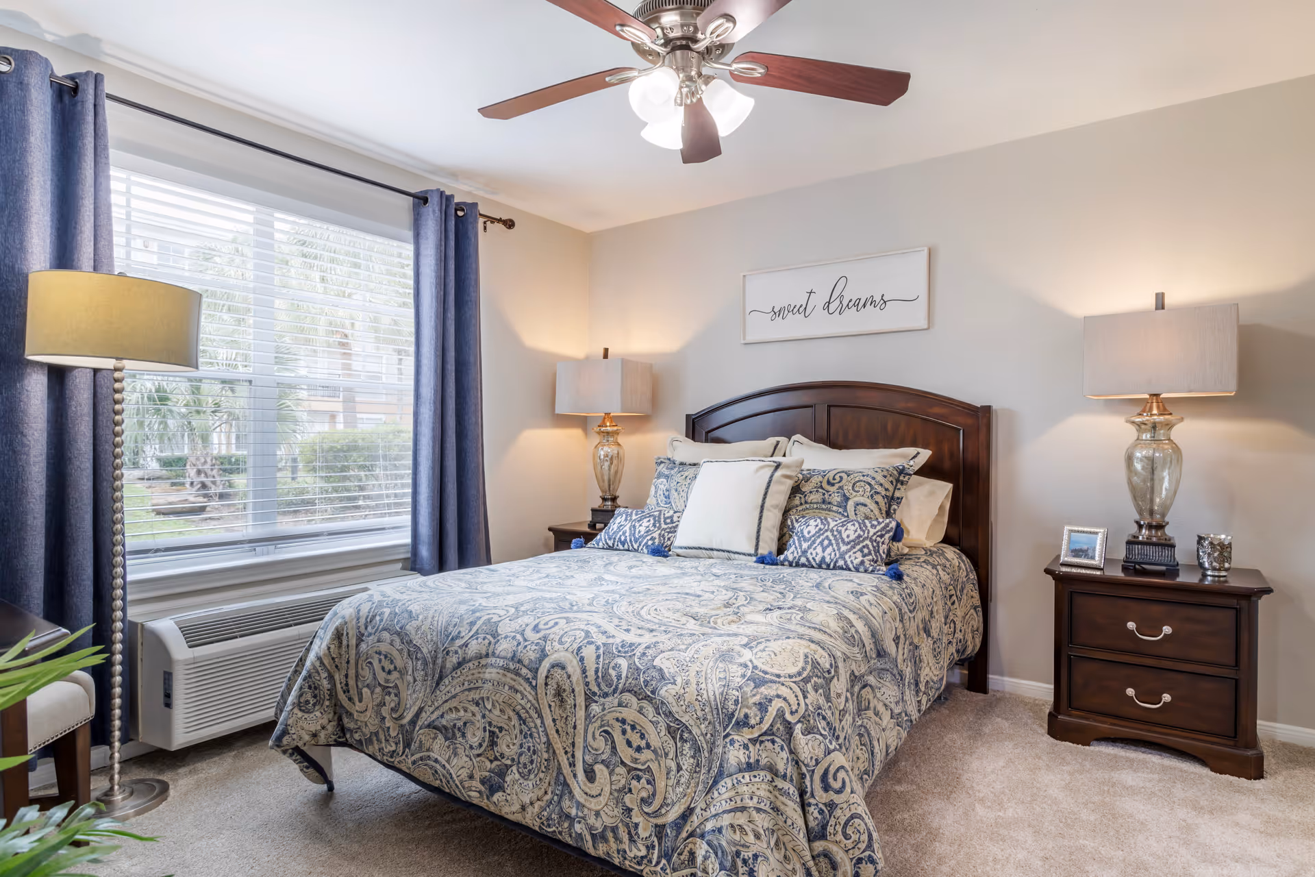 A cozy bedroom with a large bed covered in a blue and beige paisley patterned comforter and multiple pillows. The room features two wooden nightstands with matching lamps, a ceiling fan with lights, a large window with blue curtains, and a wall decoration above the bed that reads 'sweet dreams'.