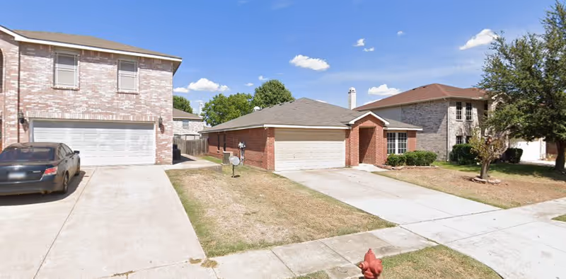 Street view of a suburban neighborhood showing three houses with driveways and garages. The house in the center is a single-story brick home with a two-car garage. The houses on either side are two-story homes with brick exteriors. There is a fire hydrant on the grass near the sidewalk in front of the center house, and a car is parked in the driveway of the house on the left. The sky is clear with a few clouds.