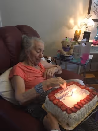 An elderly woman sitting in a recliner chair, lighting a candle on a birthday cake decorated with strawberries. A side table next to her holds a teddy bear, a lamp, greeting cards, and other small items.