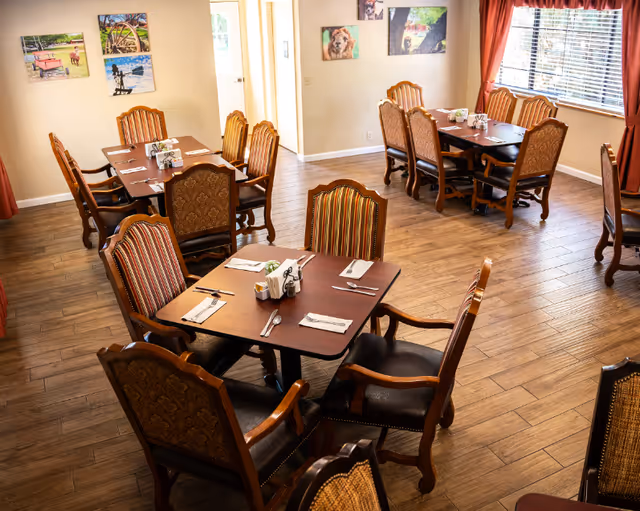 A dining room with several wooden tables and cushioned chairs arranged neatly. Each table is set with napkins, utensils, and condiments. The room has wood-patterned flooring, beige walls adorned with framed pictures, and windows with red curtains allowing natural light to enter.