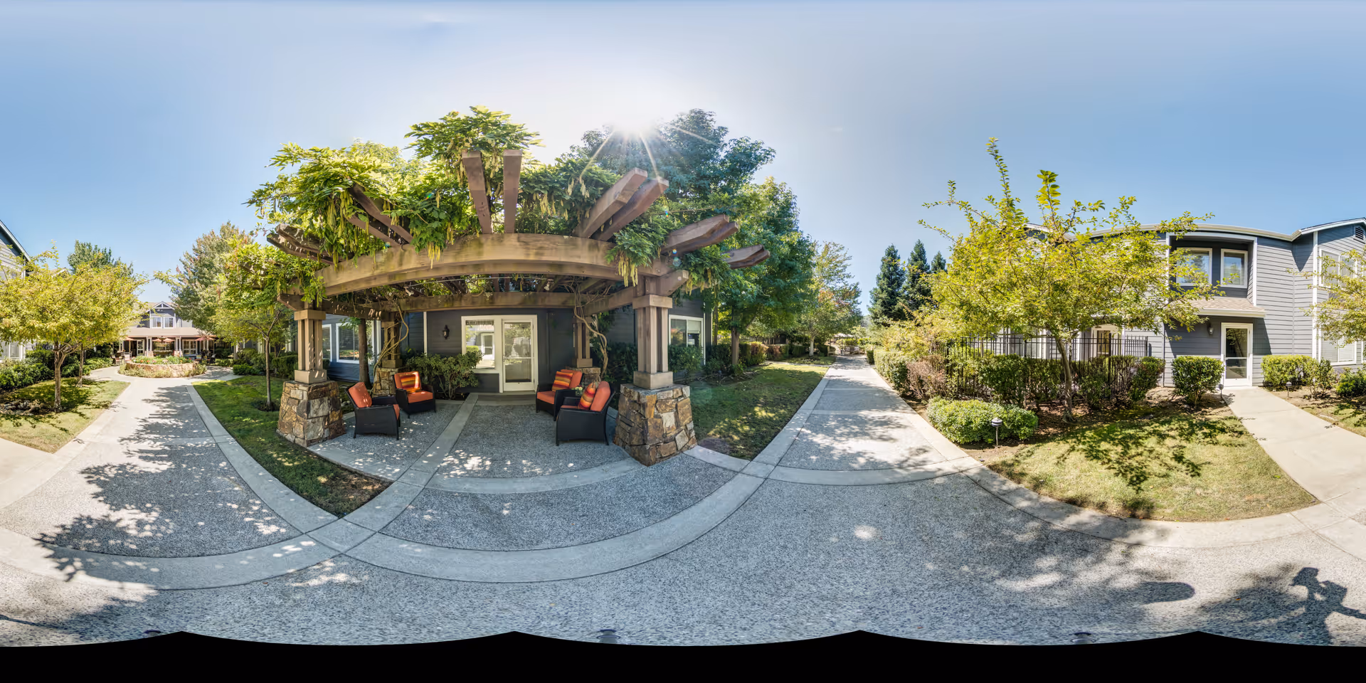 Outdoor area at Aegis Living Moraga featuring a wooden pergola with green vines and four cushioned chairs underneath. The scene includes paved walkways, green trees, shrubs, and residential-style buildings under a clear blue sky with the sun shining brightly.