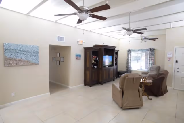A bright living room with beige tiled floors, two ceiling fans, and neutral-colored walls. The room features a large wooden entertainment center with a TV, two beige armchairs, a small wooden side table, and a window with patterned curtains. There is a hallway entrance on the left side and a white door on the right side of the room.