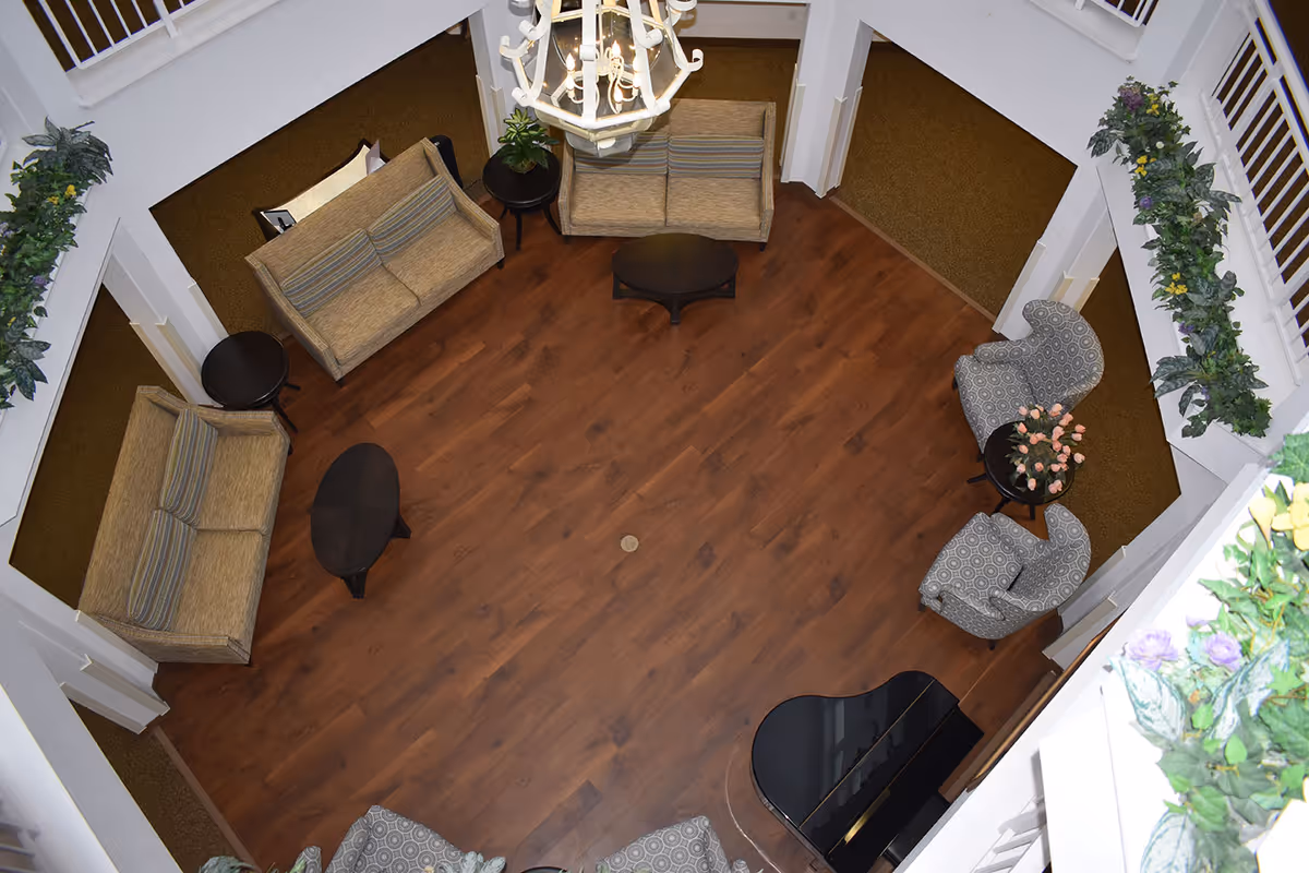 Overhead view of a senior living facility common area with wooden flooring, two beige sofas, two patterned armchairs, several small round and oval tables, a black grand piano, a chandelier, and green plants decorating the white walls.
