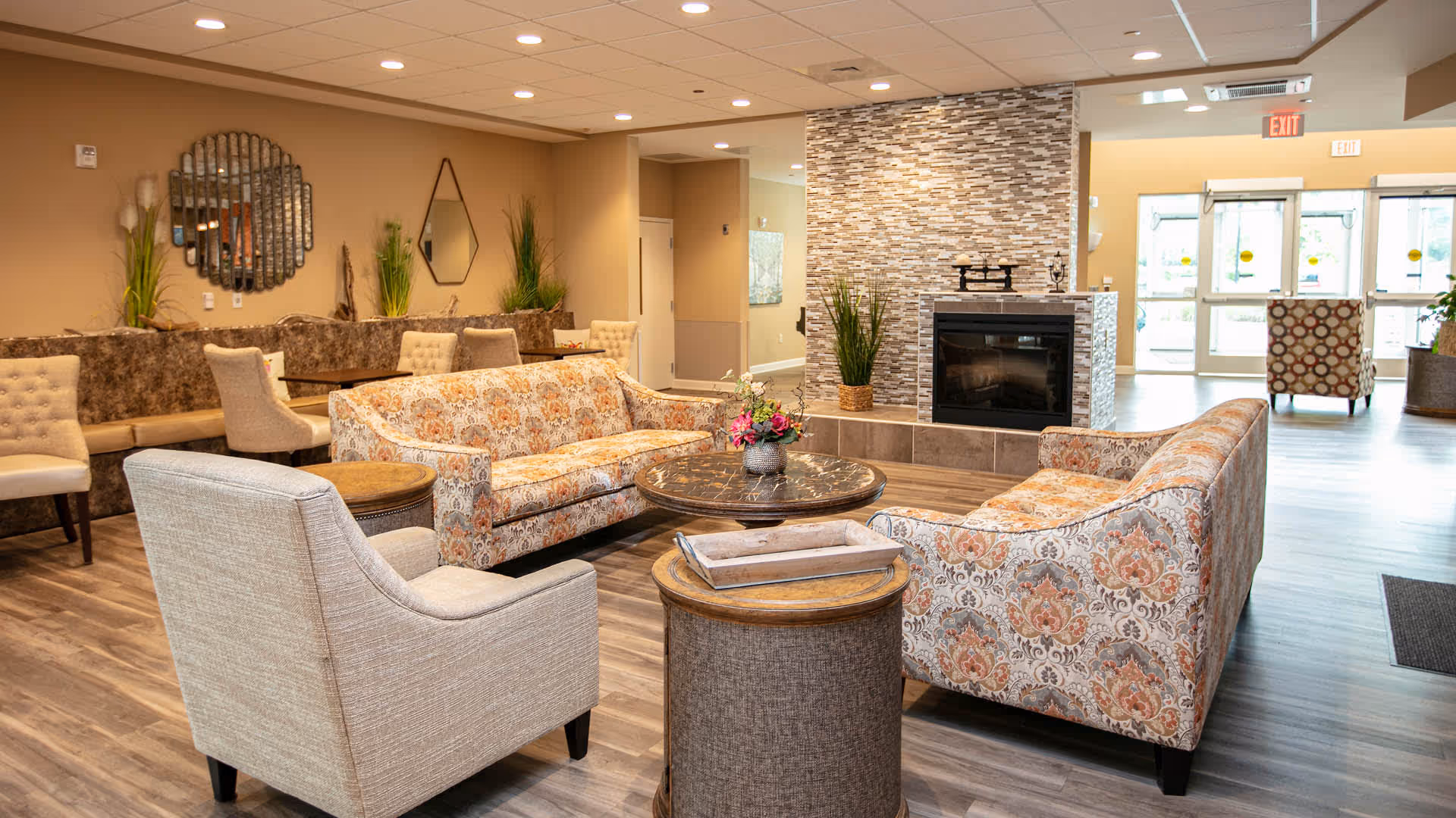 Cozy communal lounge with patterned sofas, armchairs, coffee tables, and a fireplace in a senior living facility.
