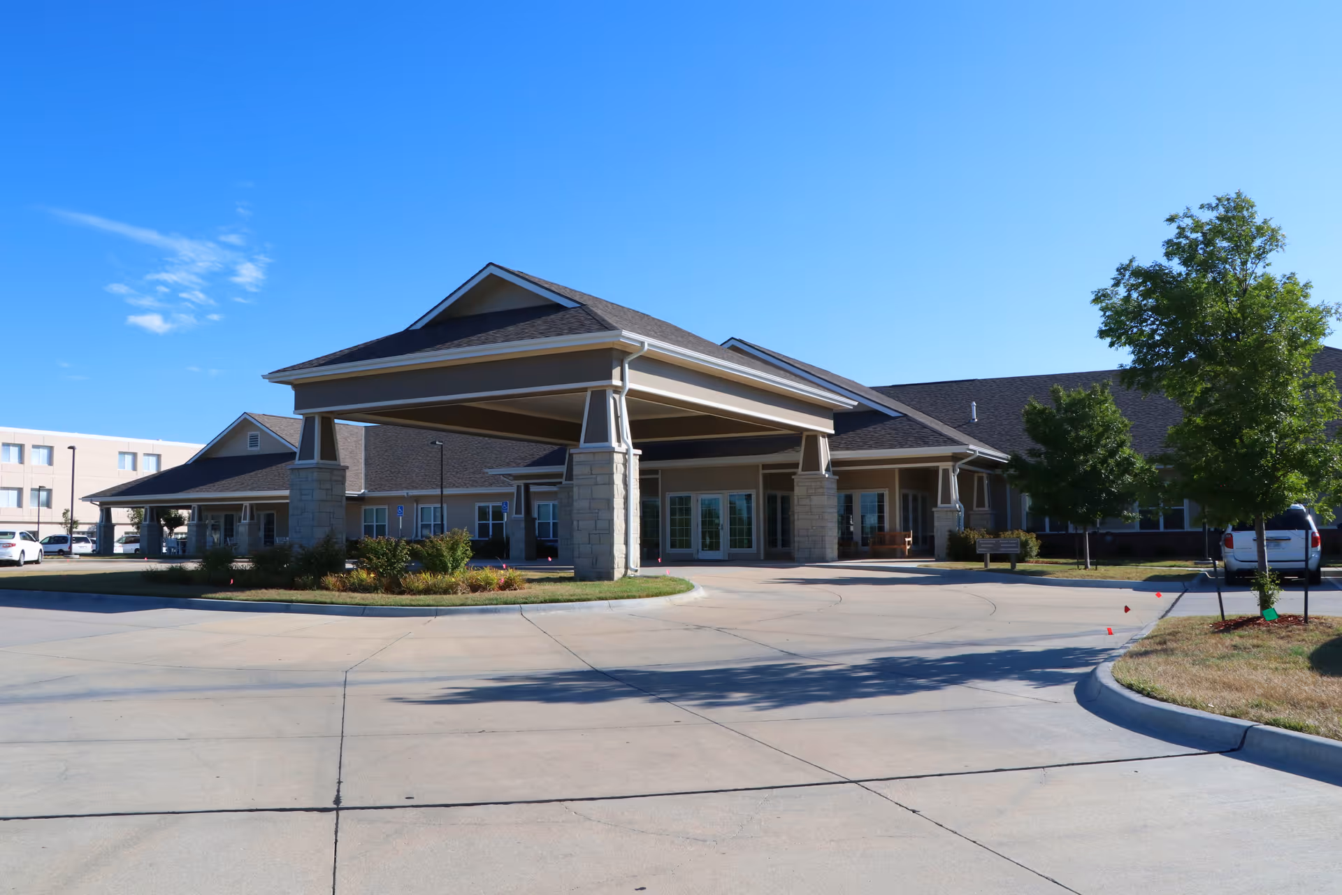 Front entrance of a single-story senior living facility with a covered porte-cochère, circular driveway, and landscaping under a clear blue sky.