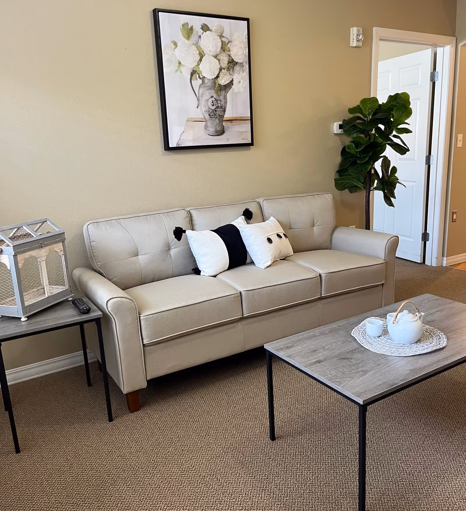 A cozy living room area featuring a beige three-seater sofa with three decorative pillows, a wooden coffee table with a white teapot and cup on a woven placemat, a side table with a decorative lantern and remote control, a potted plant in the corner, and a framed painting of white flowers hanging on the beige wall.