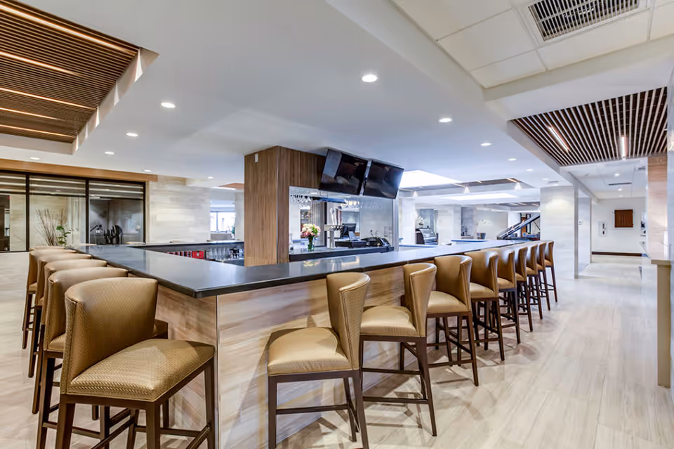 A modern indoor bar area with a long counter surrounded by high-backed bar stools. The space features light wood paneling, a black countertop, recessed lighting, and ceiling accents with wooden slats. Two flat-screen TVs are mounted above the bar, and the area is spacious and well-lit with a clean, contemporary design.