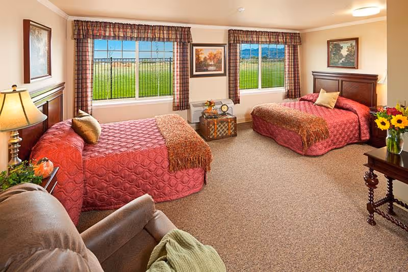 Spacious bedroom with two twin beds dressed in red quilts, a recliner, windows with plaid valances, and bedside tables.