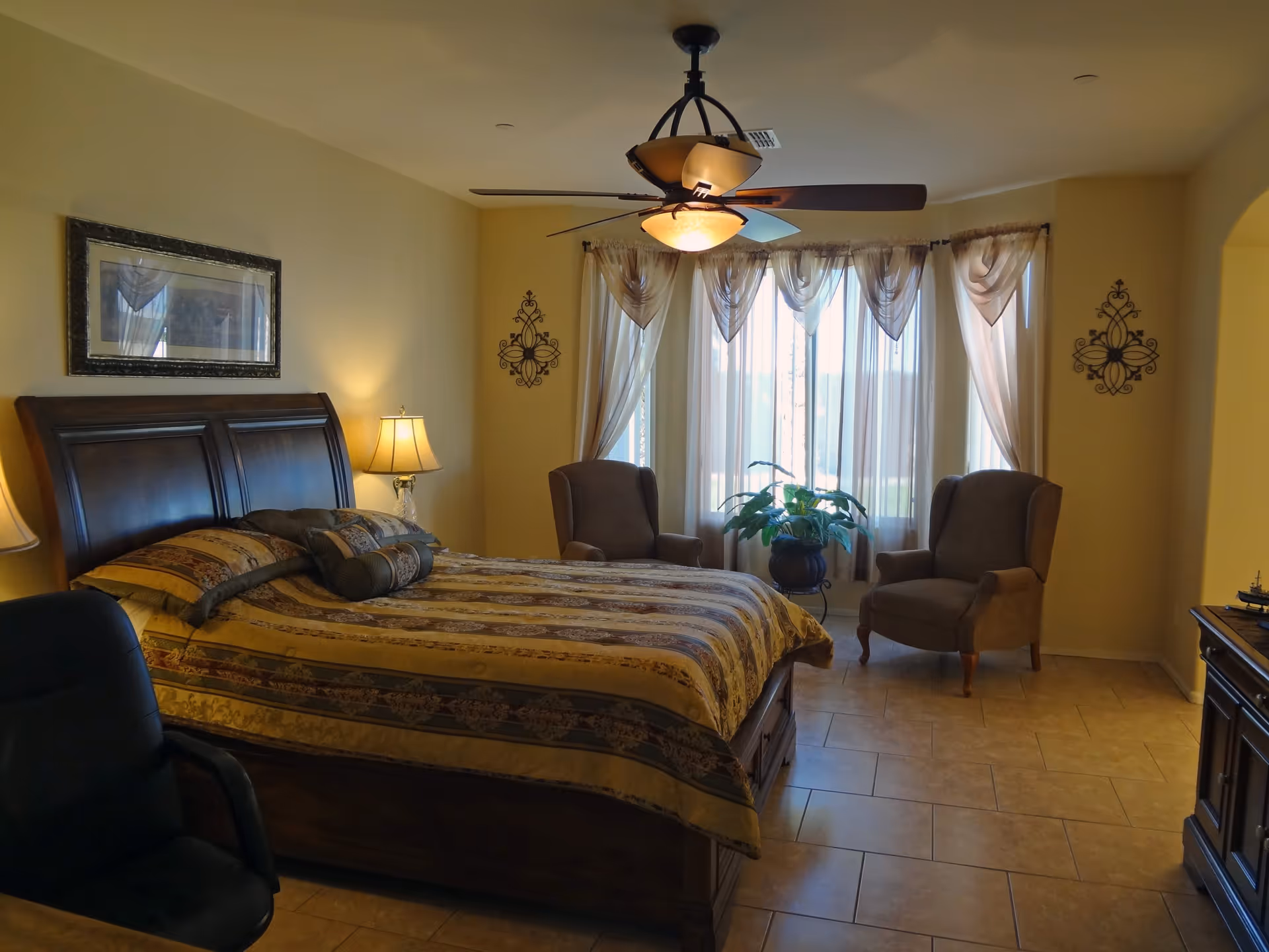 A bedroom with a large wooden bed covered with a patterned bedspread, two bedside lamps, a ceiling fan with lights, two armchairs near a window with sheer curtains, a potted plant, and a wooden dresser.