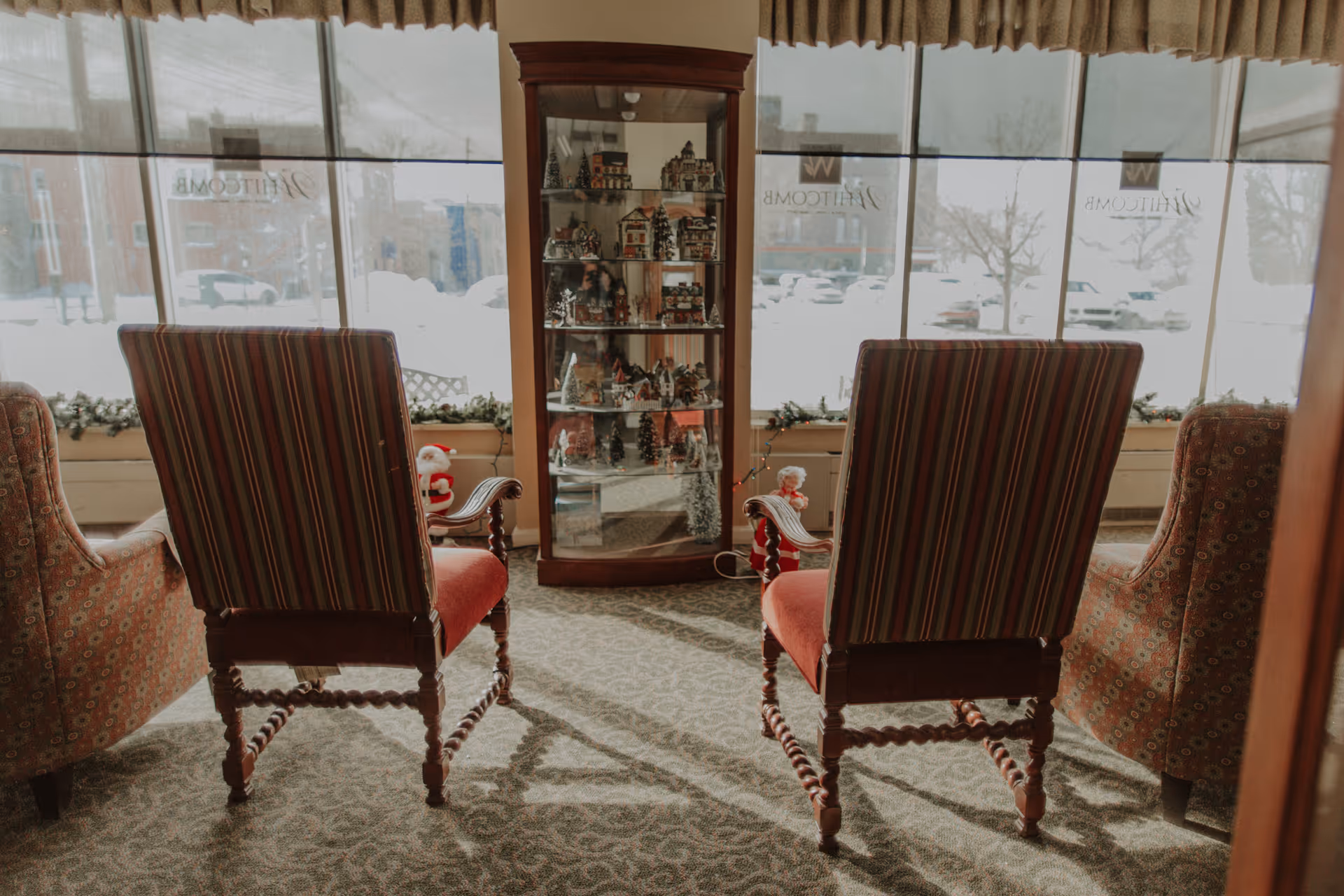 A cozy seating area in a senior living facility with two striped armchairs and two patterned armchairs arranged around a glass display cabinet filled with miniature holiday village decorations. Large windows behind the seating area show a snowy outdoor scene with parked cars and buildings. Holiday decorations, including small Santa and Mrs. Claus figures, are placed near the windows.