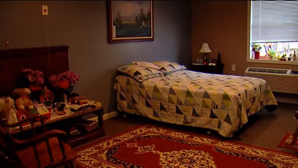 A cozy bedroom with a bed covered in a patterned quilt, a wooden headboard, a side table with a lamp, a window with blinds, and a decorative rug on the floor. There is also a small table with flowers, books, and stuffed animals.