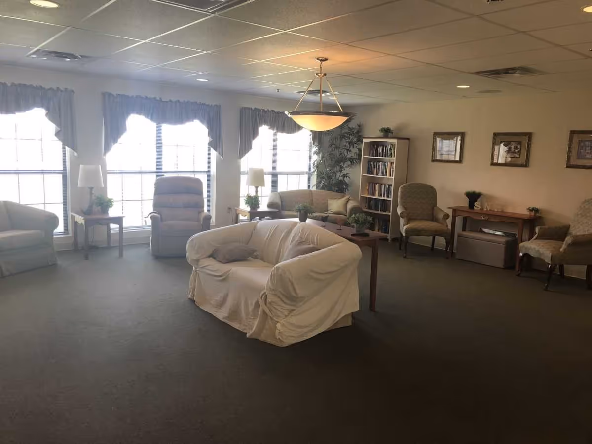 A cozy living room area with a white covered sofa in the center, surrounded by armchairs and a loveseat. There are large windows with curtains letting in natural light, two side tables with lamps and plants, a bookshelf filled with books, framed pictures on the wall, and a hanging ceiling light fixture.