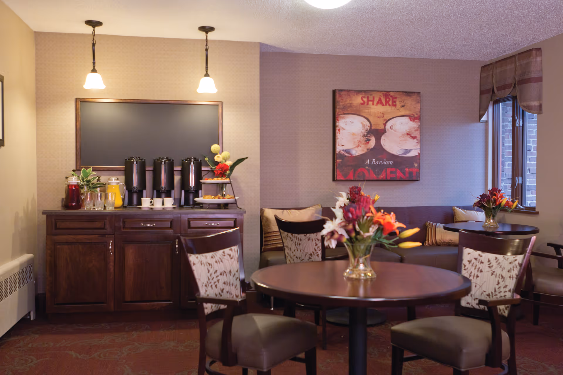 Cozy common dining area with round tables, chairs, floral centerpieces and a beverage station against the wall.