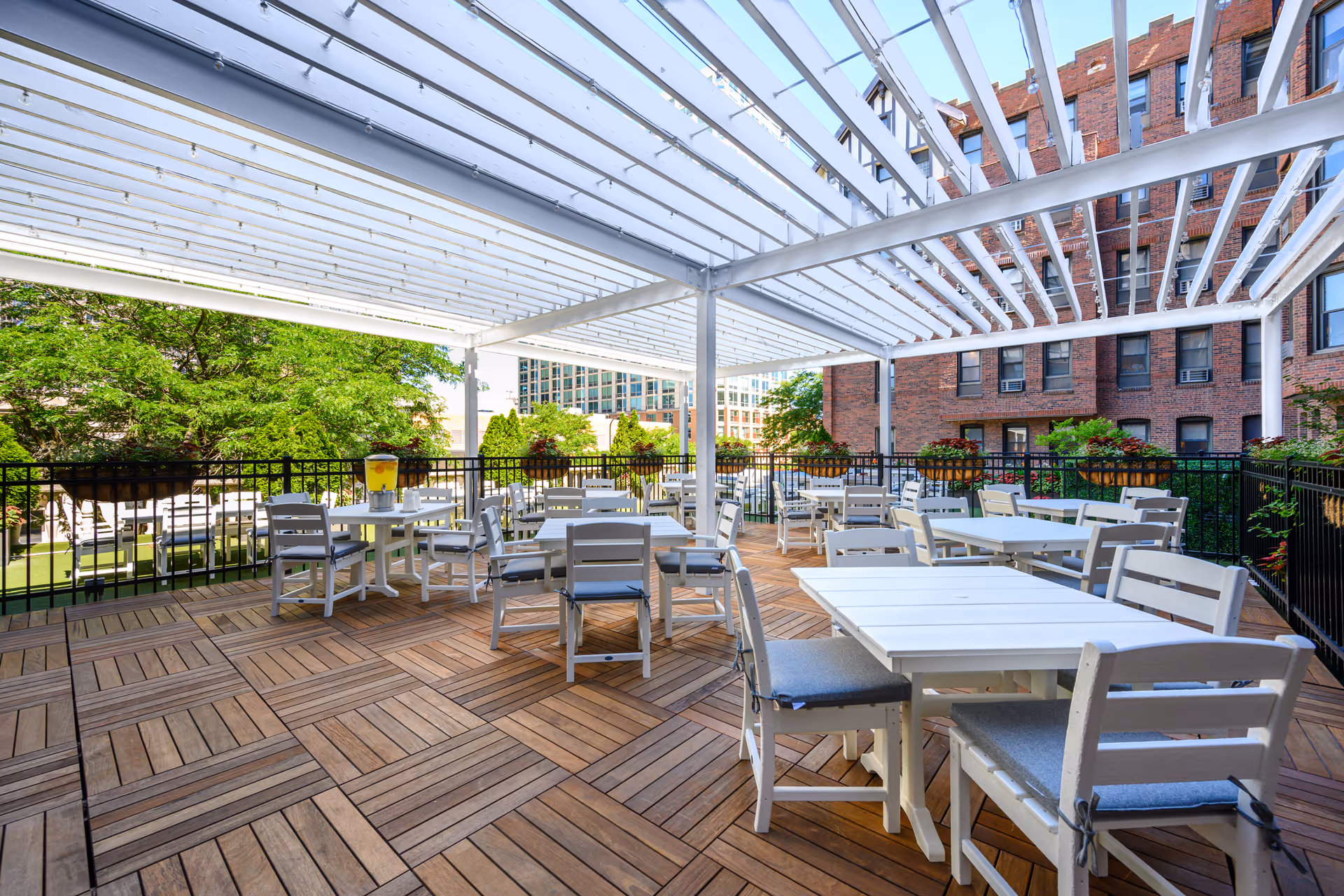 Outdoor patio area with wooden flooring and white tables and chairs under a white pergola. The patio is surrounded by a black metal railing with flower boxes and greenery. In the background, there are trees and a brick building.