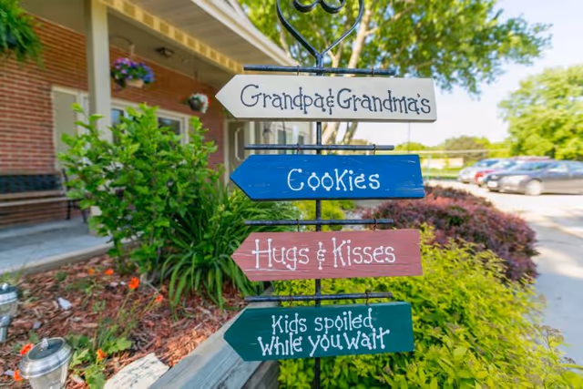 A decorative garden sign with four wooden arrows pointing in different directions. The arrows read: 'Grandpa & Grandma's', 'Cookies', 'Hugs & Kisses', and 'Kids spoiled while you wait'. The sign is placed in a landscaped garden area with green plants and flowers, next to a building with a brick wall and a parking lot with cars in the background.