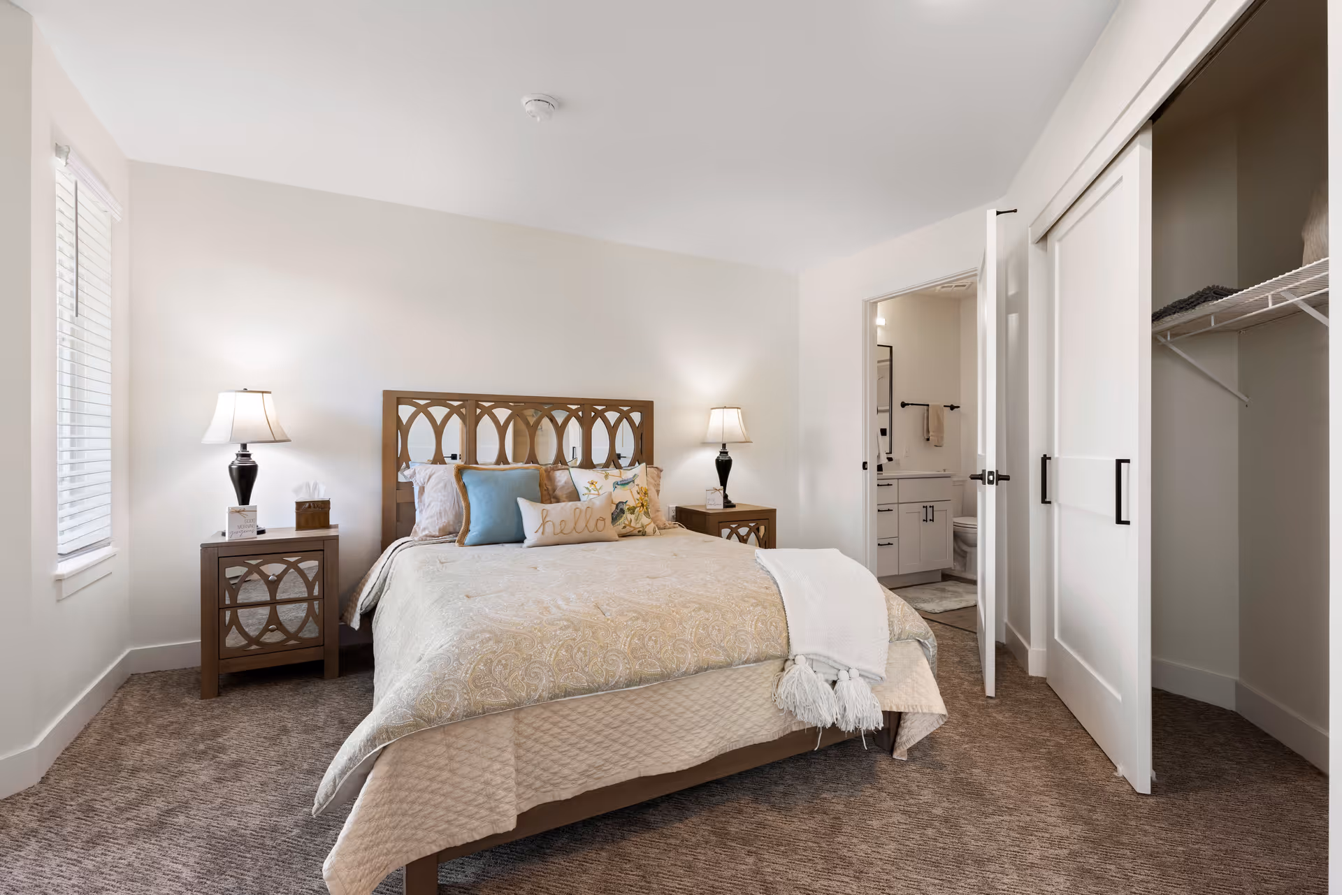 Bright furnished bedroom with a bed, matching nightstands and lamps, an open closet, and an en-suite bathroom visible through a doorway.