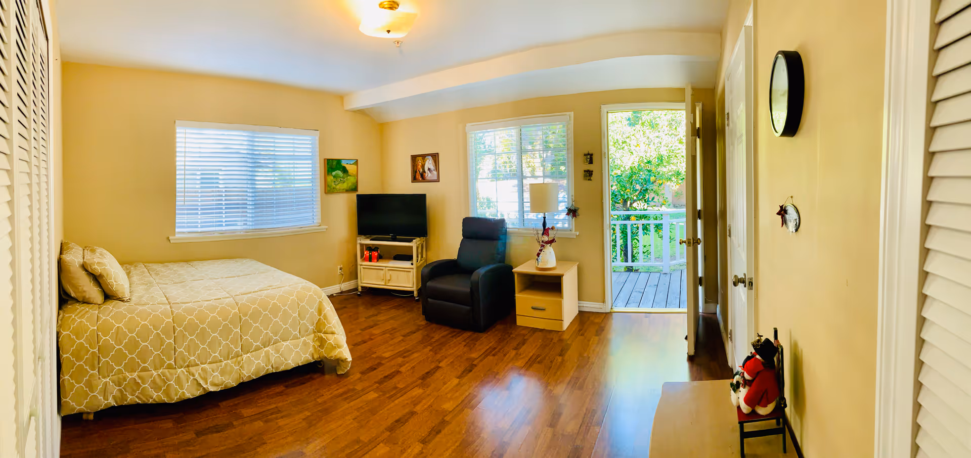 A bright and cozy bedroom with wooden flooring, a bed with a beige patterned bedspread and pillows, a black recliner chair, a small wooden nightstand with a lamp and decorative items, a TV on a stand, two windows with white blinds, and an open door leading to a wooden deck outside. The walls are painted light yellow and there is a clock on the wall.