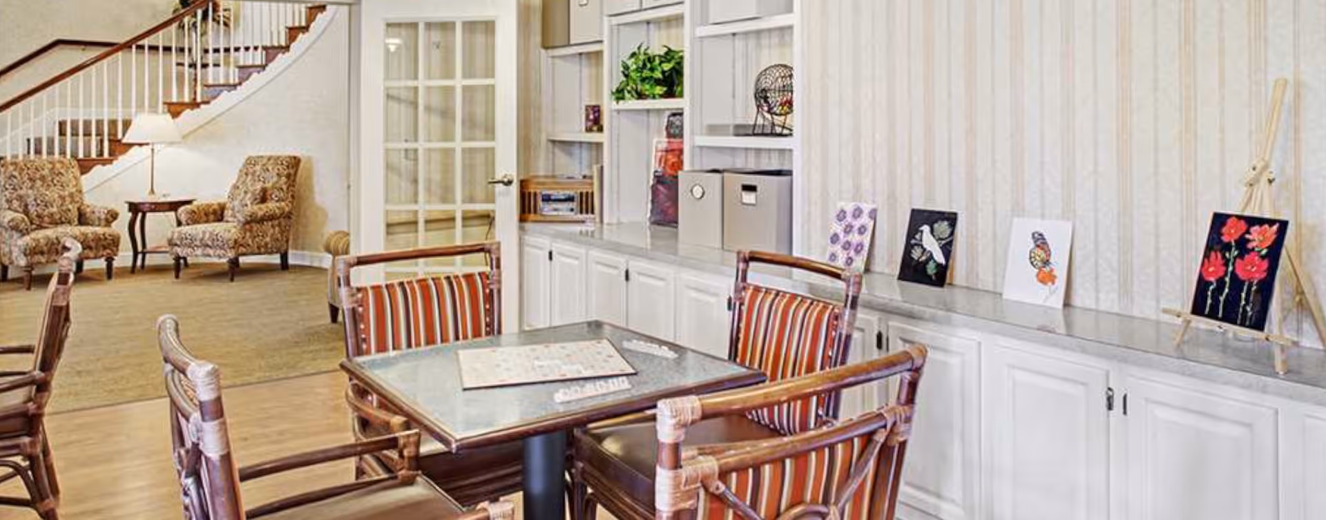 A cozy common area with a table and four striped cushioned chairs in the foreground. Behind the table is a long counter with white cabinets and several pieces of artwork displayed on top. To the left, there are two upholstered armchairs with a small table and lamp between them, and a staircase with wooden handrails leading upstairs.