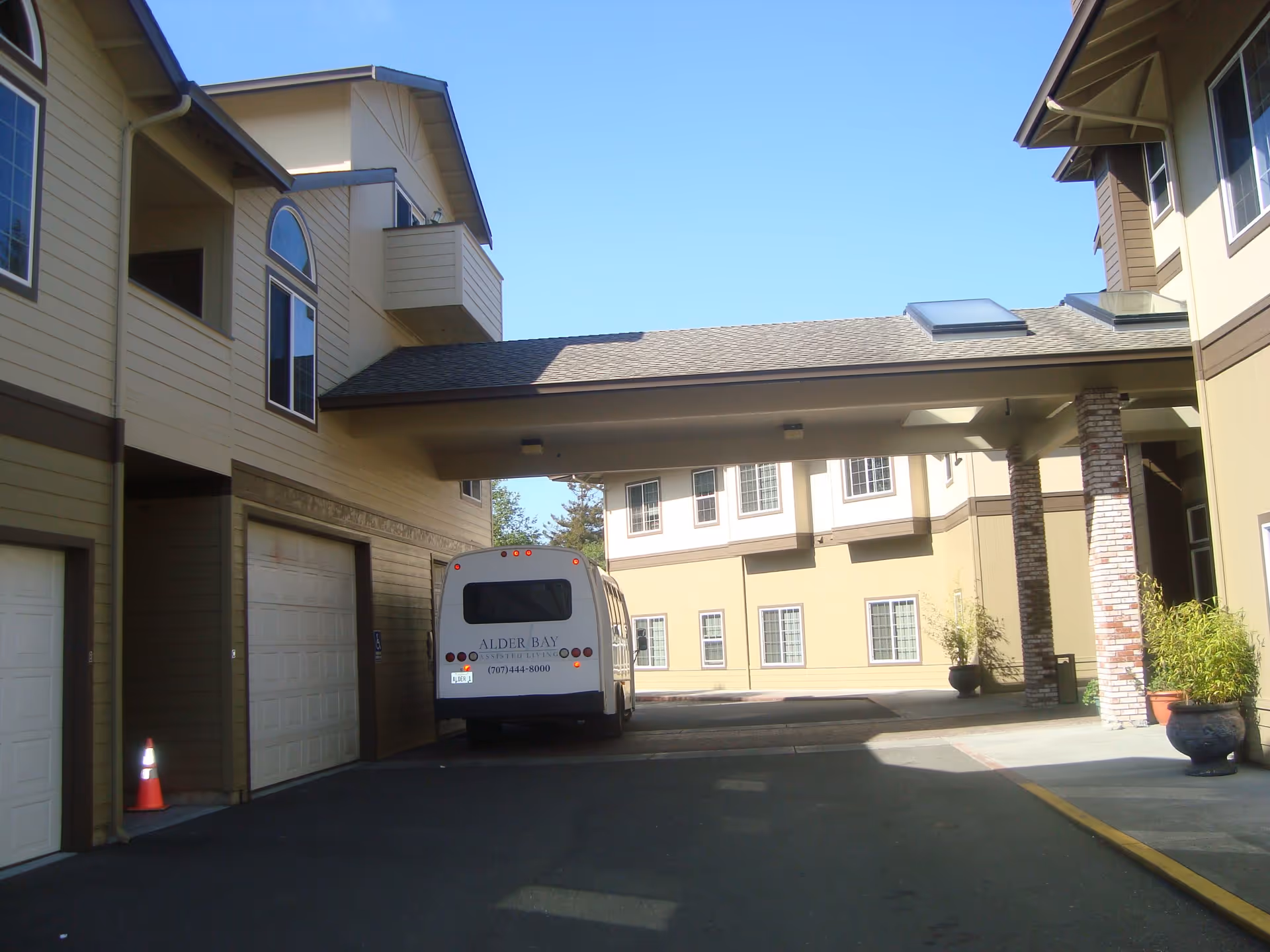 Covered driveway entrance of Alder Bay Assisted Living with a shuttle bus parked under a porte-cochère between two beige buildings.