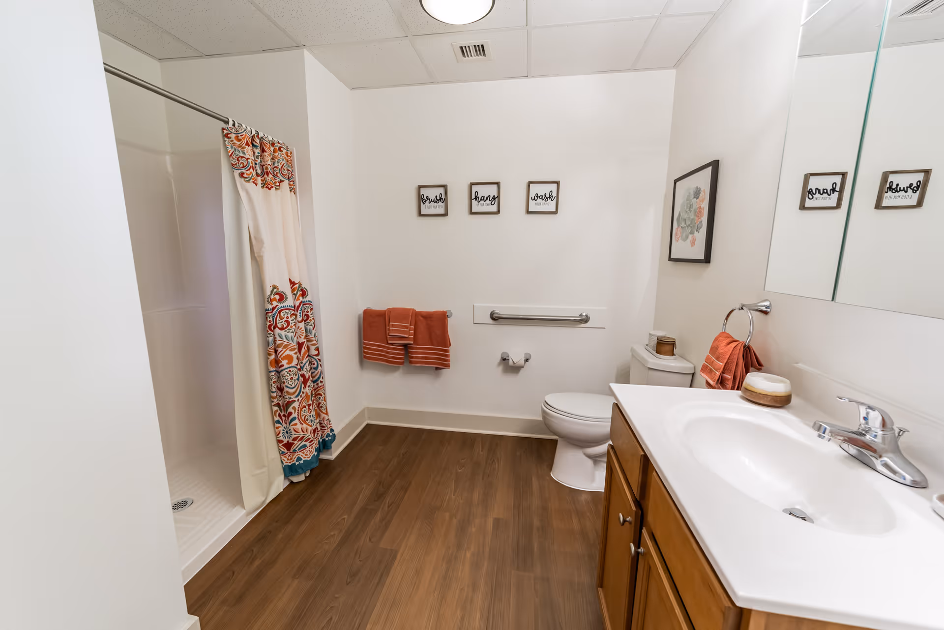 A clean and well-lit bathroom with a wooden floor, a white shower with a colorful patterned curtain, a toilet with a grab bar beside it, a wooden vanity with a white sink and faucet, a large mirror above the sink, and orange towels hanging on the wall and towel ring. Three small framed signs with the words 'brush', 'hang', and 'wash' are mounted on the wall above the toilet.