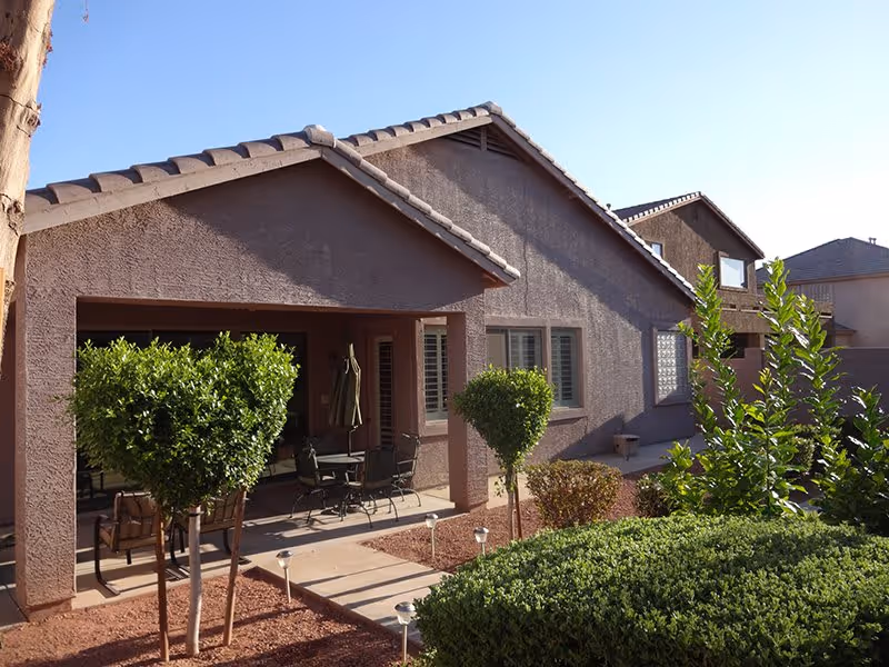 View of the exterior of a single-story residential building with a covered patio area furnished with outdoor chairs and a table. The yard features neatly trimmed bushes, small trees, and landscaping with red mulch under a clear blue sky.