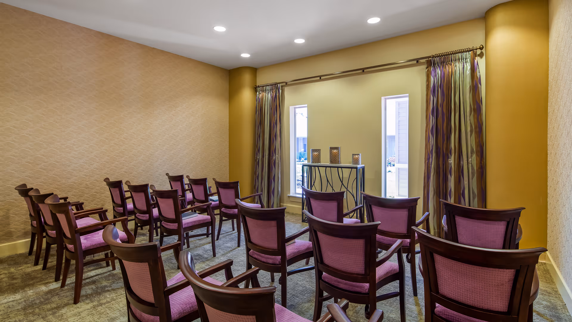 Small meeting or gathering room with rows of wooden chairs featuring purple cushions, facing a small table with three decorative candle holders. The room has beige walls, two narrow windows with patterned curtains, and carpeted flooring.