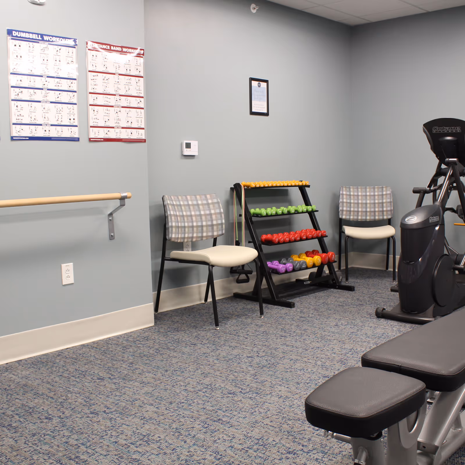 Small exercise room with a dumbbell rack, exercise equipment, chairs, and instructional posters on the wall.