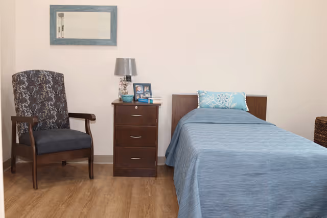 A simple senior living bedroom featuring a single bed with a blue bedspread and decorative pillow, a wooden nightstand with a lamp, framed photos, and books, a patterned armchair, and a framed mirror on the wall.