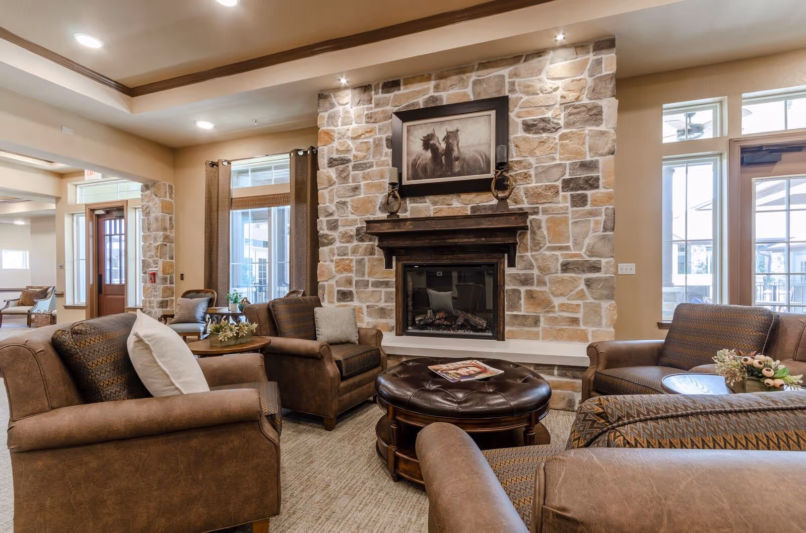 A cozy living room area with brown leather and fabric armchairs arranged around a round leather ottoman. The room features a stone fireplace with a wooden mantel, above which hangs a framed picture of two horses. Large windows with brown curtains allow natural light to enter, and there are small tables with decorative plants beside the chairs.