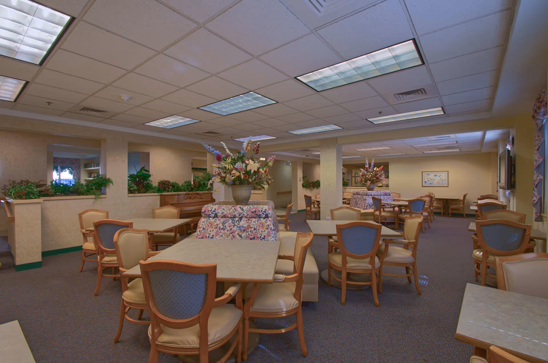 A spacious dining room in a senior living facility with multiple tables and chairs arranged neatly. The room features floral upholstered booth seating with large flower arrangements on top. The walls are light-colored, and there are several windows with floral curtains. The ceiling has recessed fluorescent lighting, and there are plants and a piano visible in the background.
