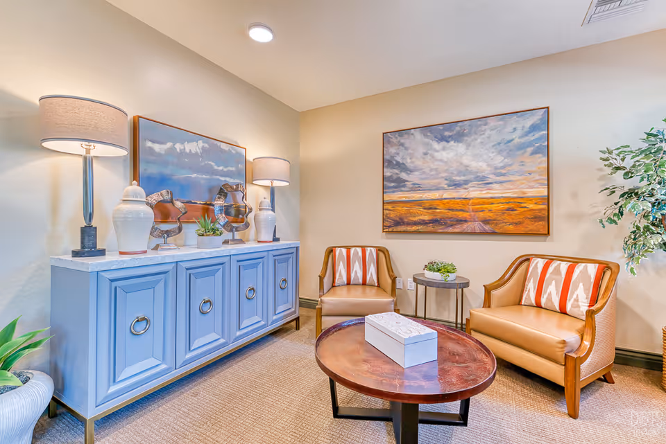 A cozy sitting area in a senior living facility featuring two tan armchairs with patterned cushions, a round wooden coffee table with a decorative box, a small side table with a plant, a blue cabinet with two table lamps and decorative items, and a large landscape painting on the wall.