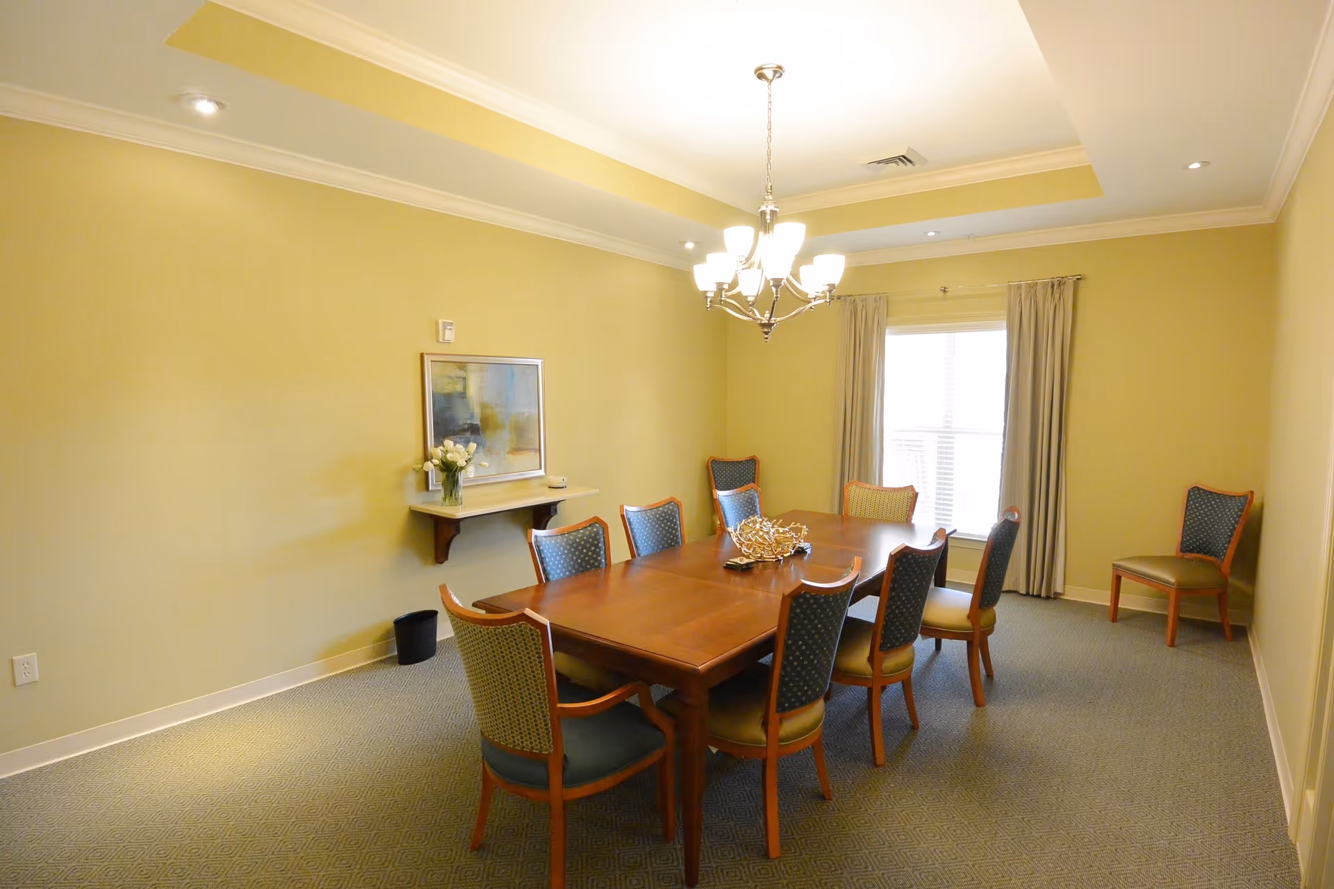 A well-lit dining room with a wooden rectangular table surrounded by eight upholstered chairs. The walls are painted light yellow, and there is a window with beige curtains letting in natural light. A chandelier hangs above the table, and a small shelf on the wall holds a vase with flowers and a framed picture.