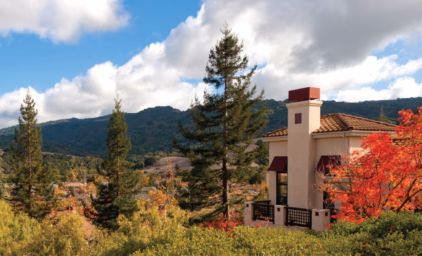 Exterior view of a beige stucco building with a red-tiled roof among autumn trees.