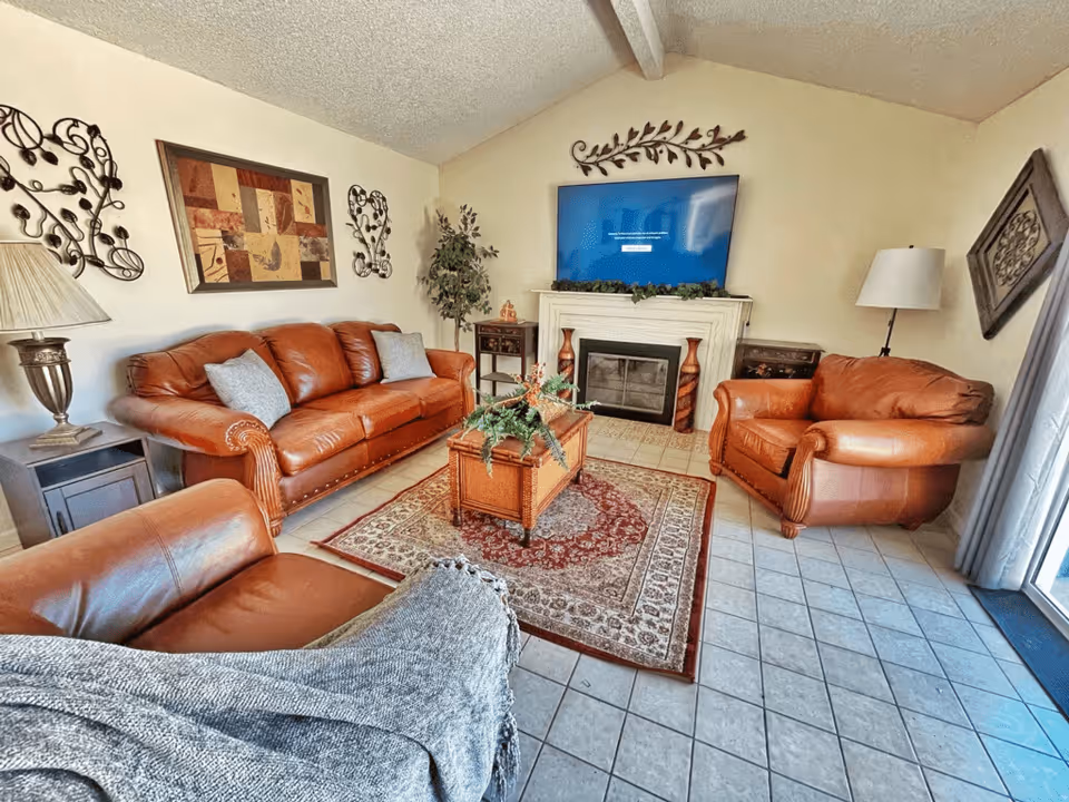Living room with brown leather sofa and armchairs, patterned rug, coffee table, fireplace with a TV, and decorative wall art.