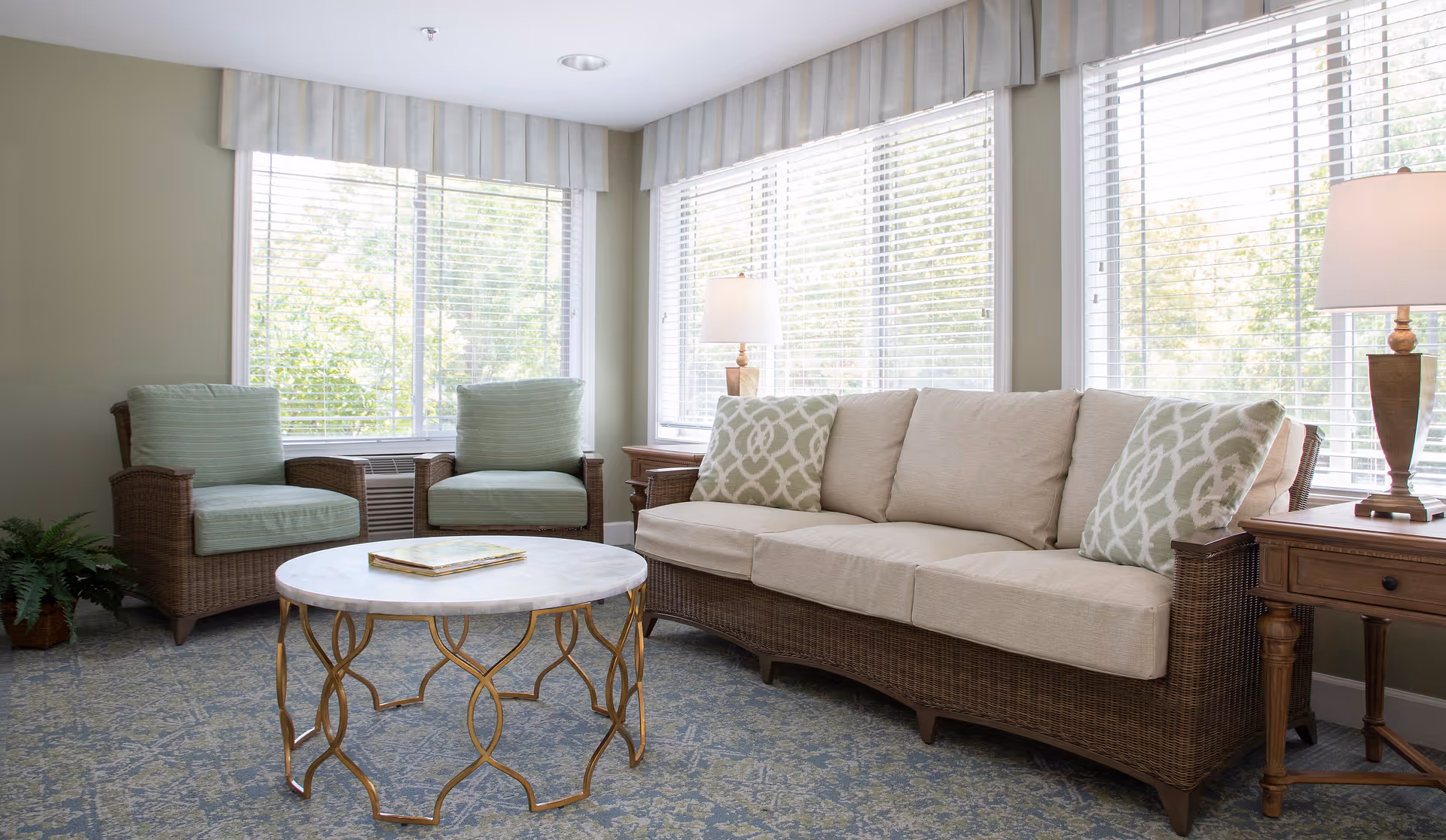 A bright and cozy living room with large windows covered by white blinds. The room features a beige cushioned sofa with patterned pillows, two matching armchairs with light green cushions, a round marble-top coffee table with a decorative gold base, two wooden side tables each with a lamp, and a potted plant in the corner.