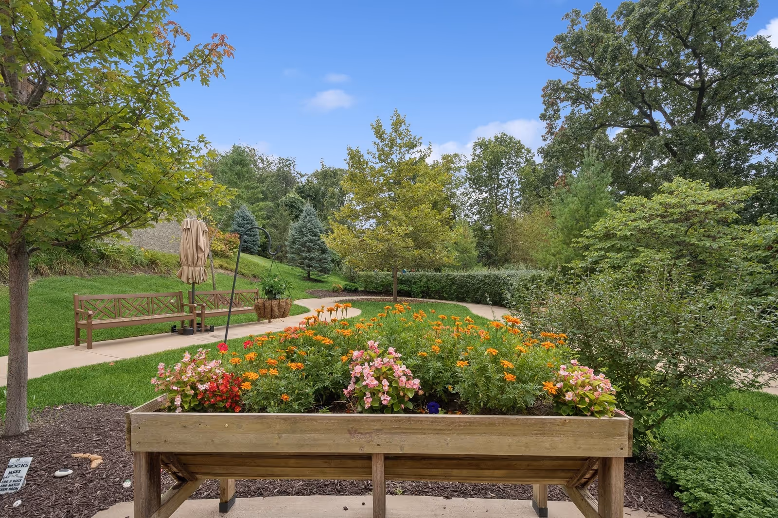 A well-maintained outdoor garden area with a raised wooden flower bed filled with colorful flowers including orange and pink blooms. There is a paved walking path winding through the green grass and trees, with wooden benches and a closed umbrella along the path. The sky is clear and blue with a few clouds.