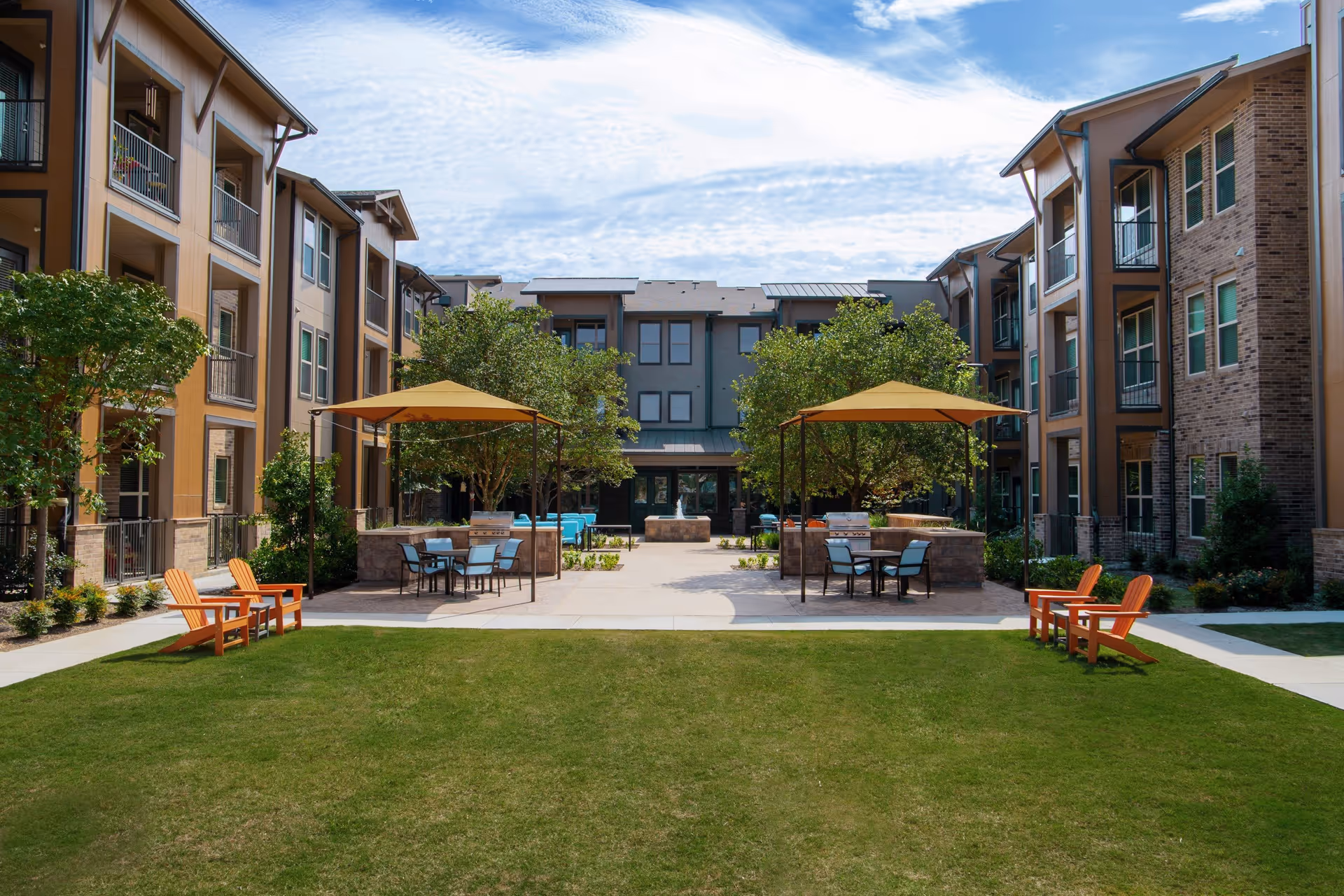 A landscaped courtyard between three-story residential buildings featuring a lawn, shaded seating areas with umbrellas, and Adirondack chairs.