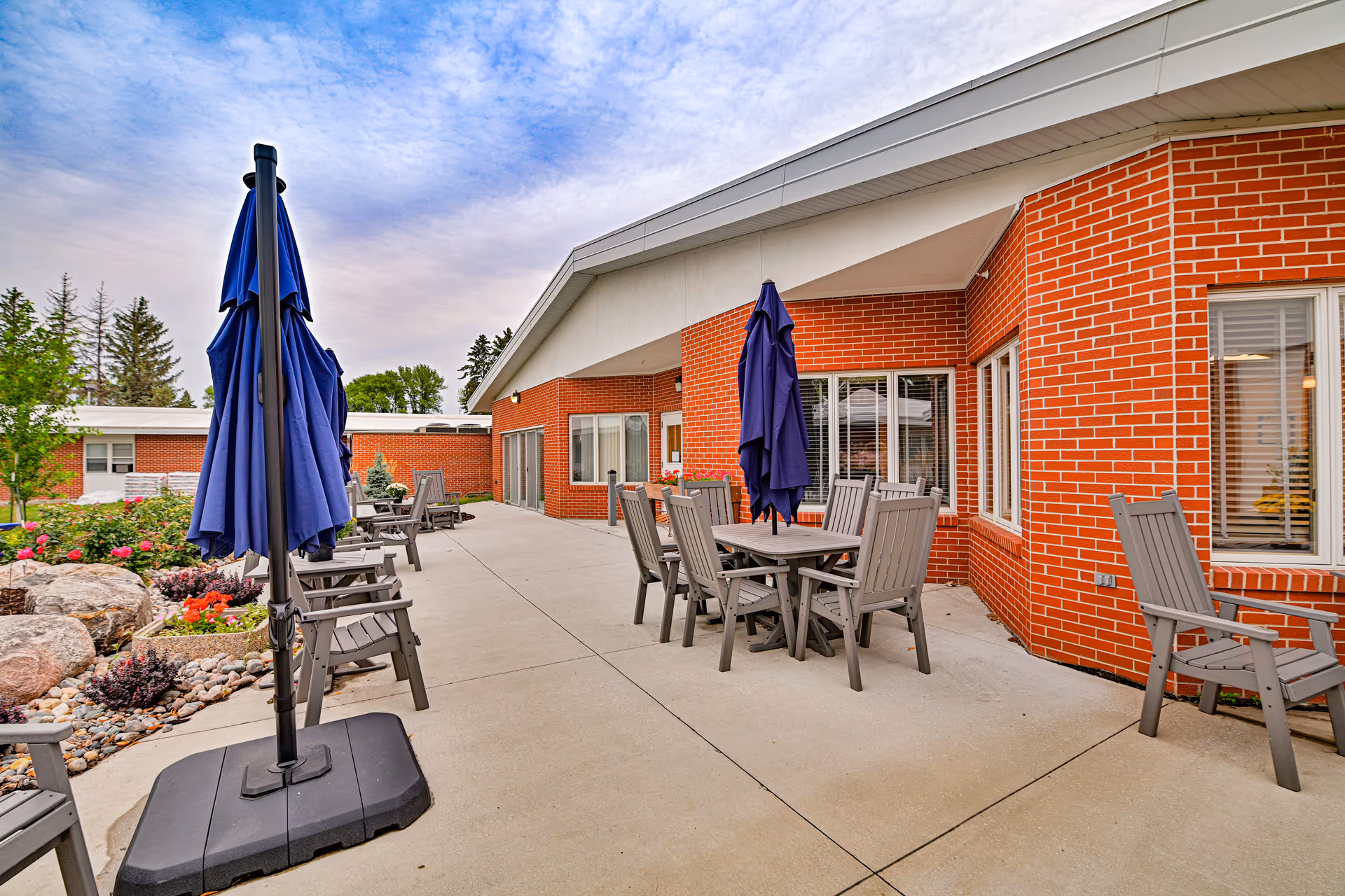 Outdoor patio area at Valley Senior Living on Cherry - Tufte Manor featuring several gray wooden tables and chairs with closed blue umbrellas. The patio is adjacent to a red brick building with multiple windows, and there are landscaped garden beds with rocks and flowers along the side.