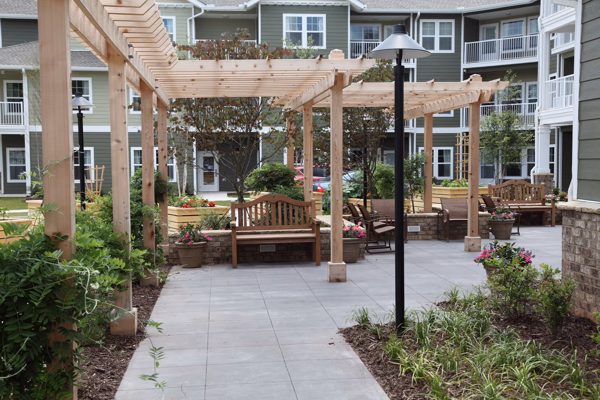 Outdoor courtyard area at Cottonwood Estates Gracious Retirement Living featuring wooden pergolas, benches, potted plants, and landscaped greenery with a multi-story residential building in the background.