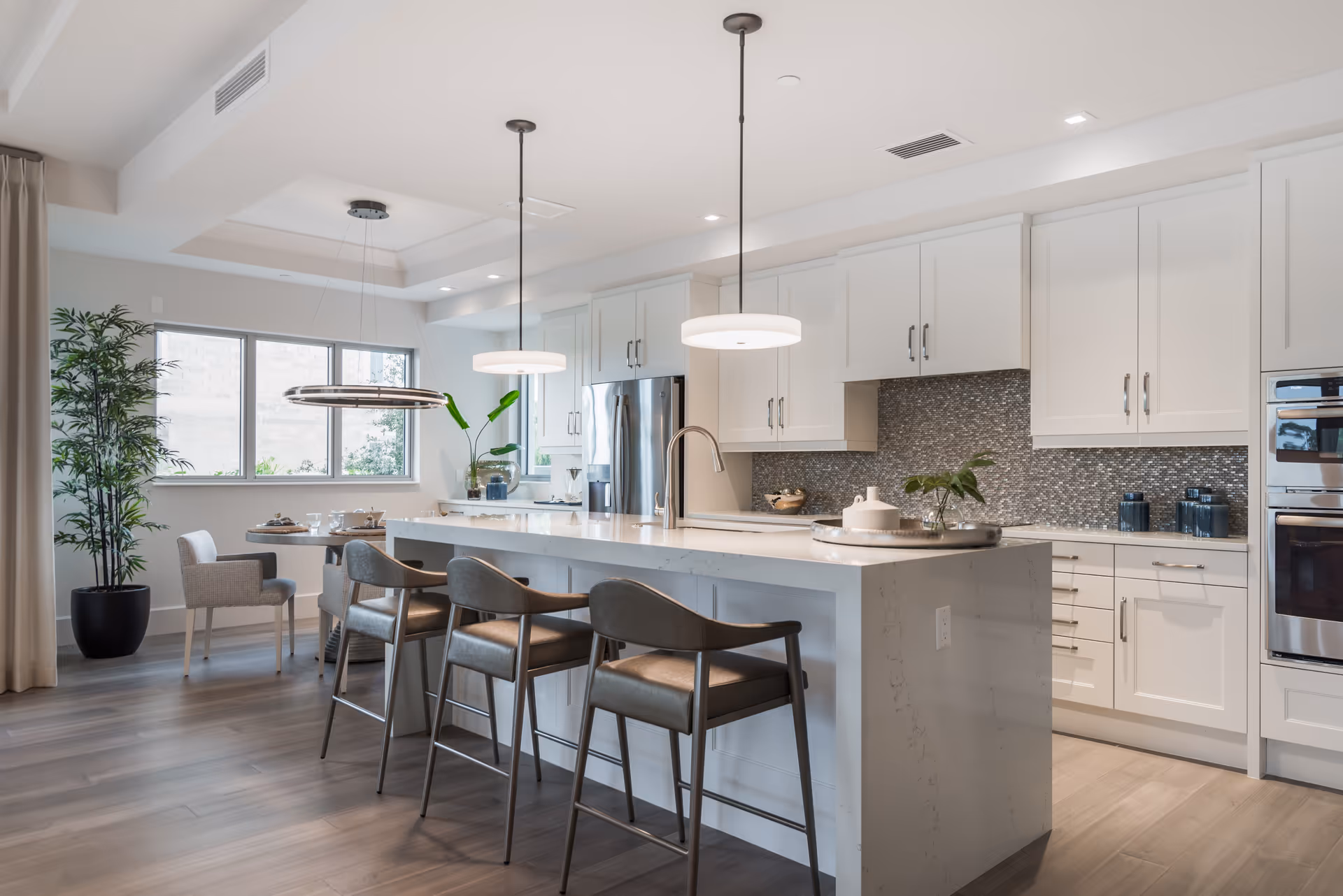 Modern kitchen with white cabinetry, a large marble island with four bar stools, two pendant lights hanging above the island, stainless steel appliances, a dining area with a round table and chairs near a window, and a tall potted plant in the corner.