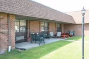 Outdoor patio area of a brick building with several chairs, a picnic table, and a lamp post on a grassy lawn.